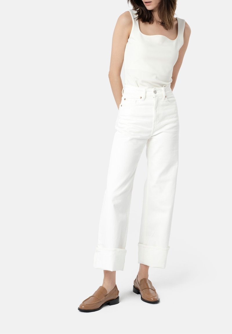 White high-waisted jeans with rolled cuffs, paired with a fitted cream tank top and brown loafers, featuring simple design and smooth texture.