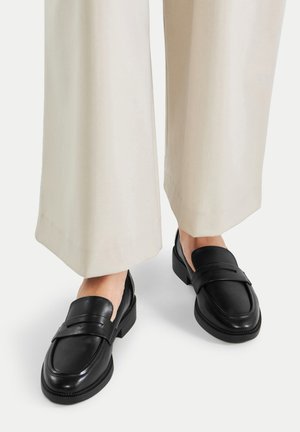 Mocasines de cuero negro con una superficie lisa, punta redondeada y una correa en la parte superior. Combinados con pantalones de pierna ancha de color claro.