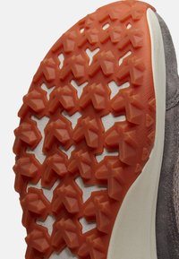 Zbliżenie podeszwy buta z pomarańczowymi gumowymi kołkami zapewniającymi przyczepność oraz białą środkową podeszwą, przedstawiające teksturowany wzór bieżnika i część szarego materiału cholewki.