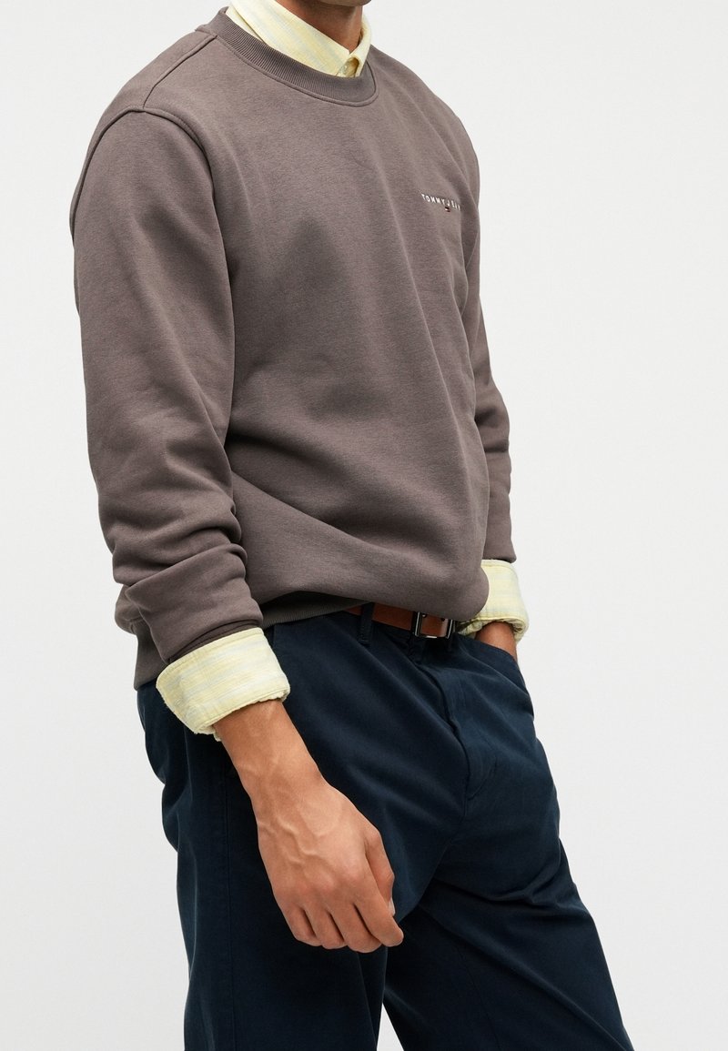 Homme portant un sweat-shirt marron col rond sur une chemise jaune avec les manches retroussées, assorti à un pantalon bleu foncé et une ceinture marron, les mains dans les poches.