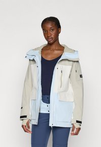 Kerge, veekindel jakk beeži ja helesinise värviga, väljasarja kapuuts, mitmed taskud ja kontrastsed tekstuurid.