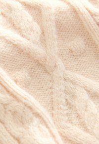 Strickschal aus weichem, hellem Beige mit strukturierten Zopfmustern und hochstehenden Noppen, der ein gemütliches, flauschiges Erscheinungsbild aufweist.