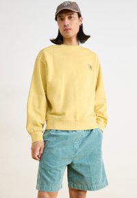 Gelbes Sweatshirt mit gerippten Bündchen und Saum, versehen mit einem kleinen Logopatch; kombiniert mit türkisfarbenen Cordshorts, die Textur und Kontrast verleihen.
