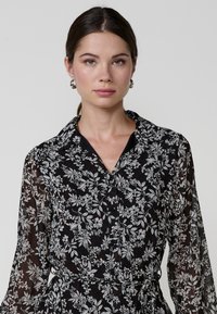 Rochie neagră cu flori, cu mâneci lungi, închidere cu butoane pe față și talie evidențiată cu un brâu. Prezintă modele florale albe elaborate pe o țesătură transparentă.