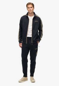 Mørkeblå track suit med guld accenter, som inkluderer en jakke med lynlås og elastiske bukser. Glat stof og logodetaljer på jakke og bukser.