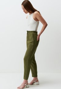 Olijfgroene high-waisted broek met plooien, taps toelopende pijpen en elastieken tailleband, gecombineerd met een witte mouwloze top en sandalen met bandjes.