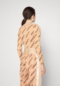 Žena s hnědými vlasy v nízkém culíku, oblečená v broskvovém volném tričku s dlouhým rukávem od Adidas a odpovídajících kalhotách s bílými bočními pruhy, stojí zády.