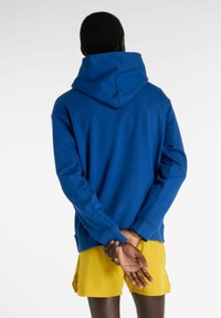 Blå huhoodtröja med avslappnad passform, ribbade muddar och fåll, mjuk textur, kombinerad med gula shorts, sedd bakifrån.