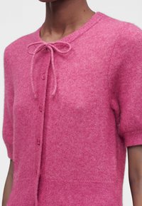 Gros plan sur une personne portant un cardigan rose duveteux à manches courtes, boutonné avec un nœud noué au niveau du col.