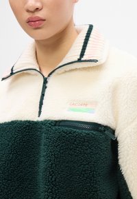 Fleece pulover s polu-zipom u krem i tamnozelenoj boji, s rebrastim ovratnikom, džepom s patentnim zatvaračem i Lacoste logotipom. Mekani, teksturirani materijal.