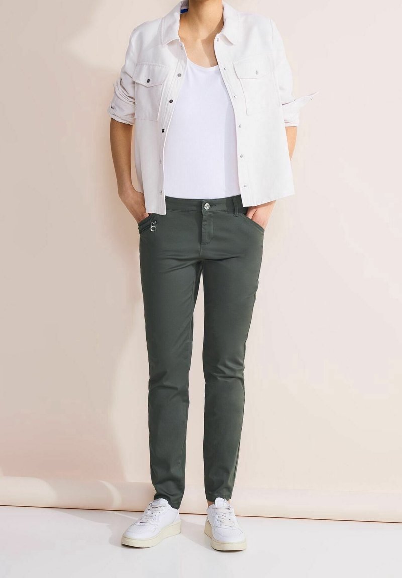 Personne portant une chemise blanche boutonnée par-dessus un débardeur blanc, un pantalon slim foncé et des baskets blanches, debout les mains dans les poches.