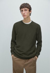 Maglione verde scuro lavorato a maglia con maniche lunghe, scollo rotondo e tessuto testurizzato, abbinato a pantaloni marrone chiaro. Design semplice.