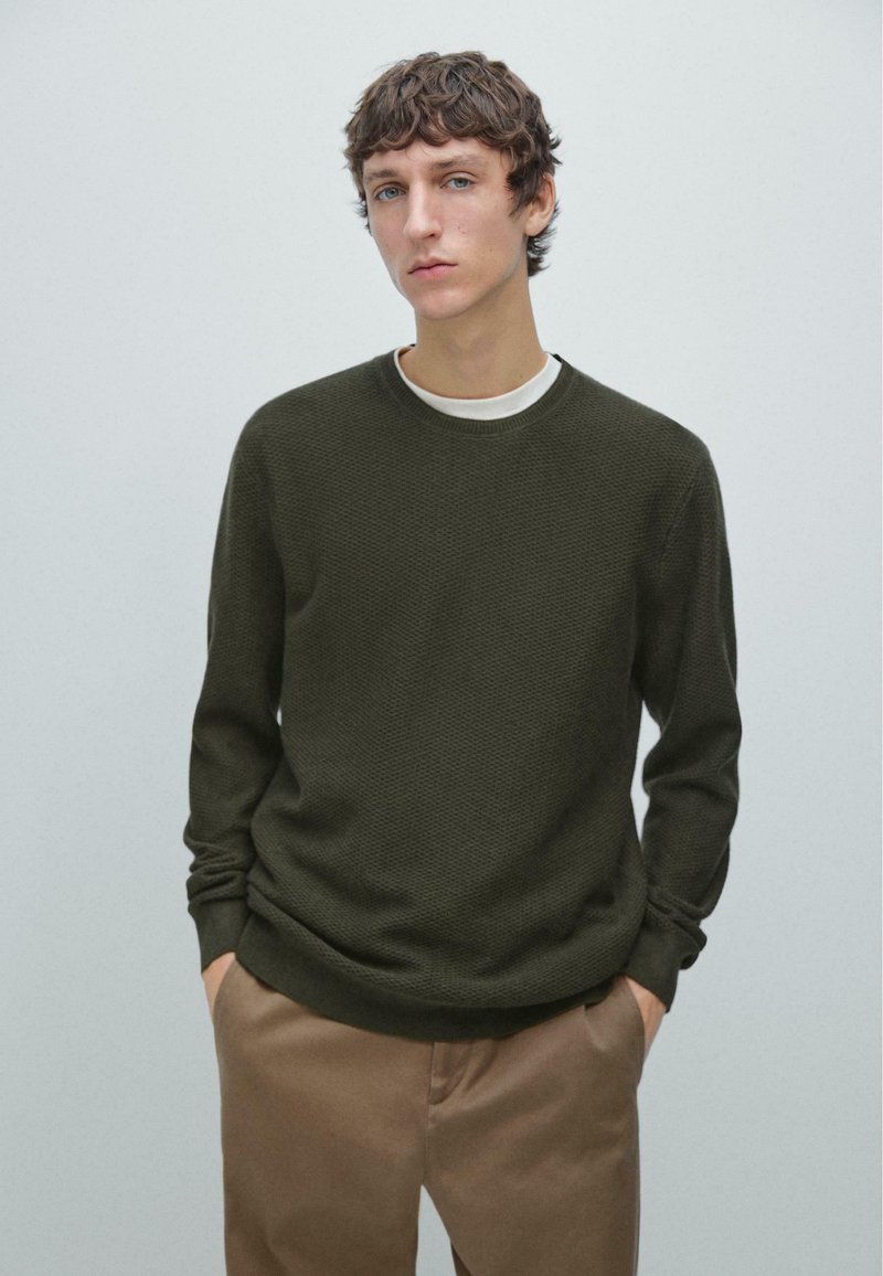 Maglione verde scuro lavorato a maglia con maniche lunghe, scollo rotondo e tessuto testurizzato, abbinato a pantaloni marrone chiaro. Design semplice.