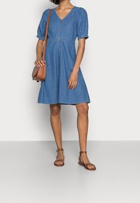 Denimmekko, jossa on lyhyet puff-hihat, V-pääntie, nappikiinnitys ja vyötärö. Yhdistetty ruskean nahkaisen olkalaukun ja sandaalien kanssa.