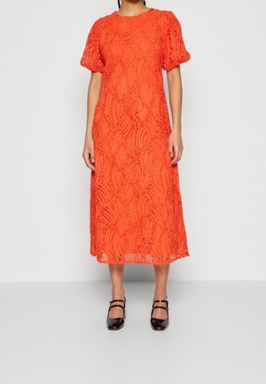 Kobieta ubrana w jasnopomarańczową sukienkę midi z bufkami i teksturowanymi wzorami spiralnymi, zestawioną z czarnymi butami na obcasie typu Mary Jane.