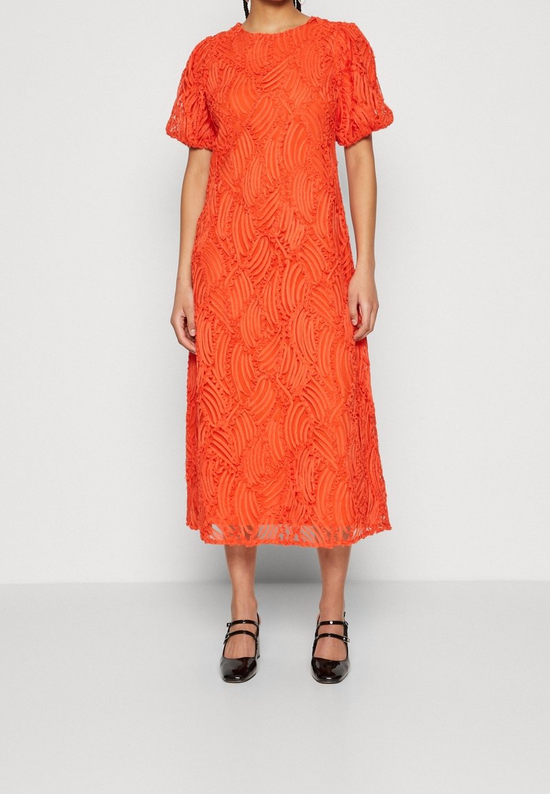 Femme portant une robe midi orange vif avec des manches bouffantes et des motifs en tourbillon texturés, associée à des escarpins Mary Jane noirs.