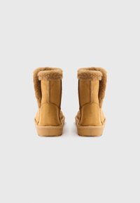 Une paire de bottes d'hiver en suède beige avec une doublure en fourrure douce repliée, vue de l'arrière sur un fond blanc uni.