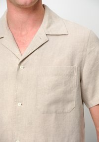 Camicia a maniche corte color beige chiaro con colletto rilassato, taschino frontale e una texture sottile. Presenta bottoni bianchi e un design casual.