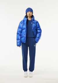 Blaue Pufferjacke mit glänzender Oberfläche, kombiniert mit einem passenden blauen Hemd und marineblauen Sweatpants. Weiße Sneaker vervollständigen das Outfit.