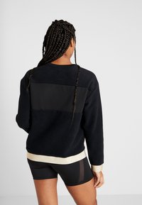 Musta fleece huppari, jossa on kontrastivärinen tekstuuri-paneeli ja kultainen metalliviimeistely helmassa. Lyhyet hihat ja rento malli.