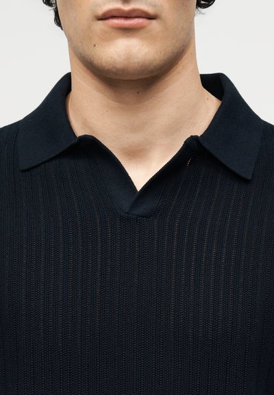 Κοντινό πλάνο του κάτω μέρους του προσώπου και του λαιμού ενός ατόμου που φοράει μαύρο πλεκτό πουλόβερ με μαλακό γιακά και V-λαιμόκοψη.