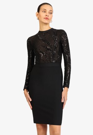 Frau trägt ein schwarzes Kleid mit einem Pailletten-verzierten, gemusterten, durchsichtigen Langarmoberteil und einem einfarbigen, eng anliegenden Rock, steht mit den Händen an den Seiten.