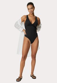 Maillot de bain noir une pièce avec un décolleté en V profond, un design en torsion à l'avant et des jambes hautes ; associé à un couvre-maillot blanc transparent et des sandales noires.