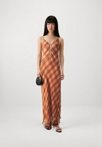 Woman with long black hair wearing a sleeveless orange plaid maxi dress with cutouts, holding a small black handbag and wearing black strappy heels.