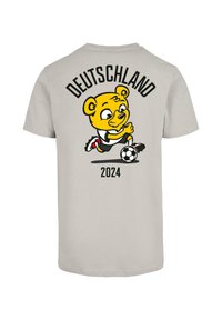Pilkas marškinėliai su žaismingu geltonu animaciniu lokiu, po užrašu "VOKIEČIAI 2024", su futbolo kamuolio akcentu.