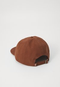 Brun bomull caps med en buet skjerm, synlig søm, og en metalljusterbar stropp bak. Teksturet stoff med ventilasjons hull.