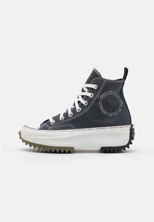 Baskets montantes en toile noire avec lacets blancs, semelle blanche épaisse avec texte, et semelle extérieure rainurée verte et noire.