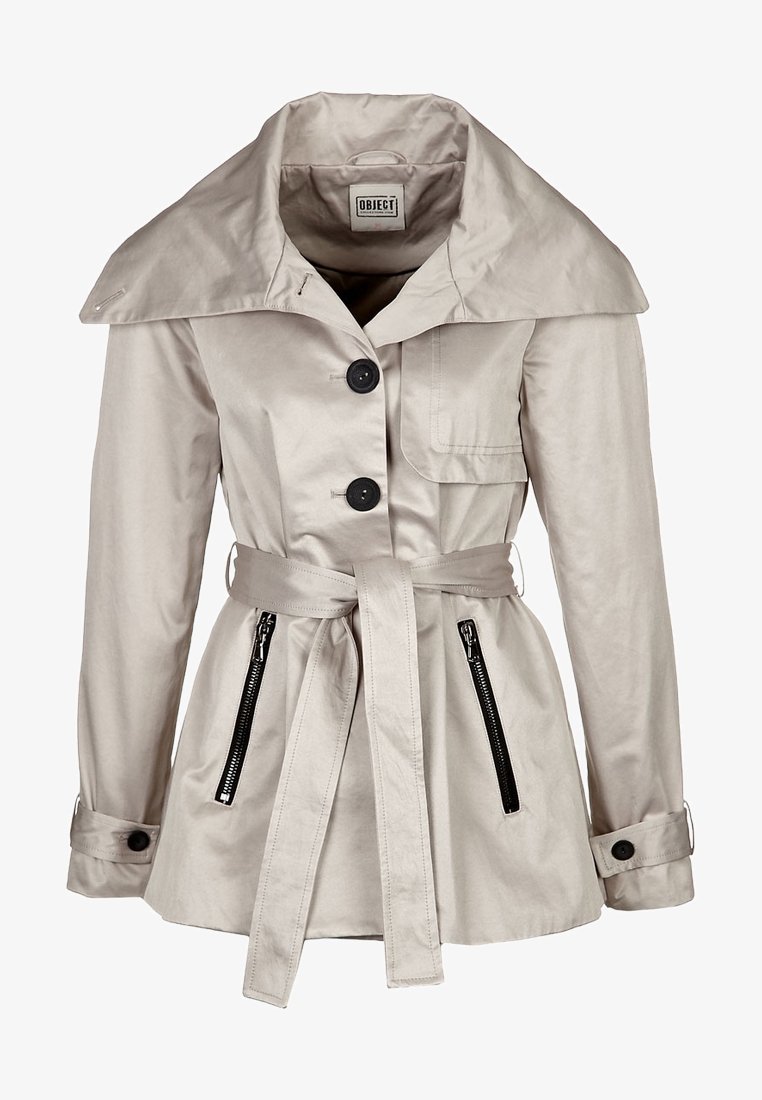 Trench beige con un amplio collar, botones negros, cintura ceñida con cinturón y bolsillos con cremallera. Textura suave y silueta ajustada.