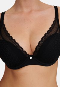 Svart spetsbh med genomskinliga paneler och vågiga kanter. Har ett geometriskt mönster och en liten metallisk detalj i mitten av bygeln.