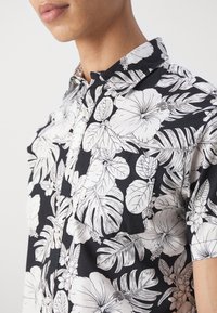 Camicia a maniche corte con sfondo nero e grandi motivi floreali e fogliari bianchi. Presenta un colletto botton-down e una tasca sul petto.