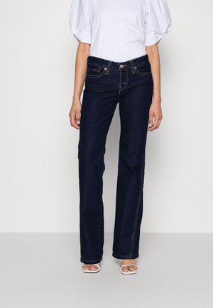 Kvinde iført mørkeblå straight-leg jeans, hvid top med pufærmer og hvide åbne hæle, står foran en ensfarvet lys baggrund.