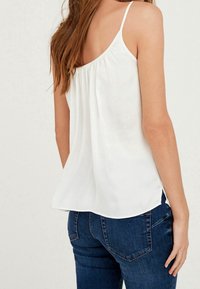 Hvid ærmeløs top med ruched rygdetalje og tynde stropper, parret med tætsiddende blå jeans. Blød, glat tekstur.
