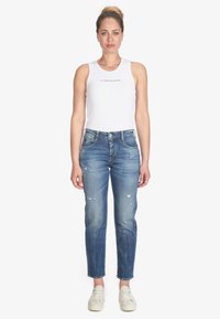 Canottiera bianca a coste con logo stampato, abbinata a jeans distressed azzurri chiari con orli sfrangiati e leggeri sbiadimenti, indossati con sneakers bianche.