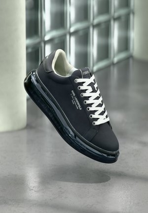 Zapatilla negra Karl Lagerfeld con cordones blancos, logo visible en el lateral y suela transparente, suspendida sobre un suelo de hormigón pulido.