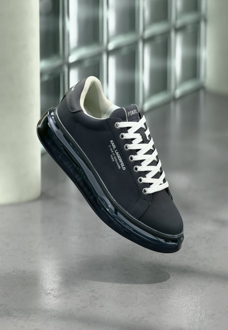 Fekete Karl Lagerfeld sneaker fehér fűzőkkel, oldalán jól látható logóval és átlátszó talppal, polírozott betonpadló felett lebegve.