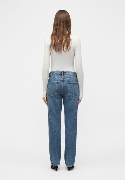 Γυναίκα με μακριά καστανά μαλλιά, ντυμένη με λευκό εφαρμοστό μακρυμάνικο τοπ και μπλε τζιν παντελόνι ψηλόμεσο, όρθια με την πλάτη προς το μέρος μας, μπροστά σε ένα απλό φόντο.