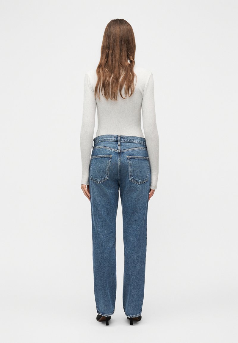 Femme aux longs cheveux bruns portant un haut blanc ajusté à manches longues et un jean bleu taille haute, debout, tournée de dos sur un fond uni.