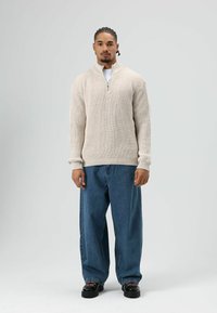 Jonge man met gevlochten haar, gekleed in een beige gebreide trui, losse blauwe spijkerbroek en zwarte schoenen, staand tegen een effen lichte achtergrond.