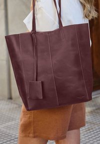 Borsa a spalla in pelle bordeaux con due manici lunghi, design piatto e un piccolo tag attaccato. Texture liscia con dettagli di cucitura visibili.