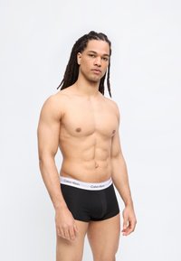 Άνδρας χωρίς πουκάμισο με μακριά dreadlocks, φορώντας μαύρα μποξέρ Calvin Klein, στέκεται μπροστά σε λευκό, απλό φόντο.