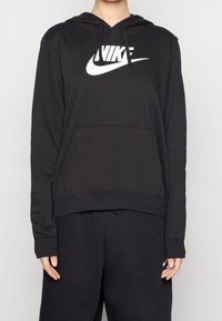 Person som bär en svart Nike-hoodie med vit logotyp och matchande svarta sweatpants, händerna avslappnat vid sidorna, enkel ljusbakgrund.