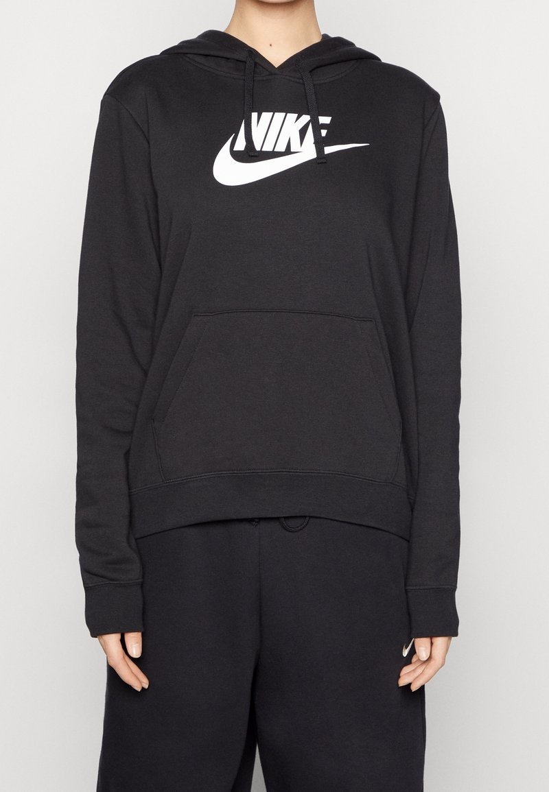 Person som bär en svart Nike-hoodie med vit logotyp och matchande svarta sweatpants, händerna avslappnat vid sidorna, enkel ljusbakgrund.