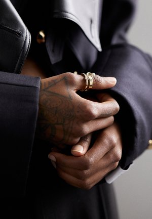 Gold rings worn on a hand with intricate designs, holding another hand. The background features dark fabric textures in black tones.