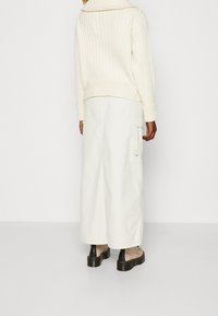 Krémový žebrovaný svetr s vysokým límcem, kombinovaný s dlouhou bílou sukní s velkými kapsami a béžovými botami s černými detaily.