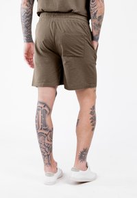 Shorts en tissu brun avec une taille élastique et une poche arrière, portés avec des baskets blanches, révélant des tatouages détaillés sur les jambes.