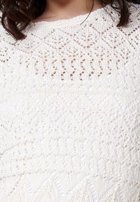 Bílý pletený svetr s průsvitnými vzory, zdobený texturovaným designem diamantů a geometrických tvarů. Kulatý výstřih a krátké rukávy.